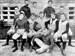 A team of eleven men posing in early football uniforms in two rows, in front of a brick building
